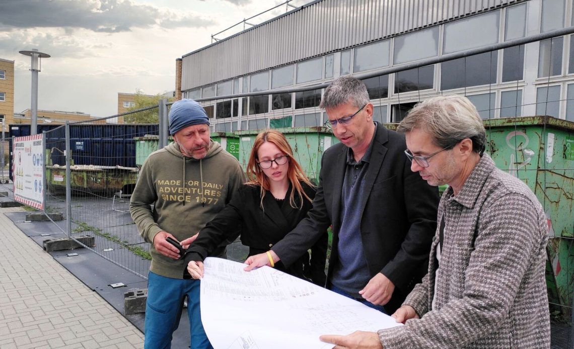 Eine Gruppe von einer Frau und drei Männern halten einen Bauplan in den Händen und begutachten ihn vor einem von einem Bauzaun abgesperrten Gebäude.