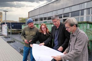 Eine Gruppe von einer Frau und drei Männern halten einen Bauplan in den Händen und begutachten ihn vor einem von einem Bauzaun abgesperrten Gebäude.