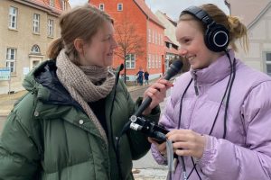 Zwei junge Studentinnen stehen auf der Straße und interviewen sich mit einem Aufnahmegerät und einem Mikrofon.