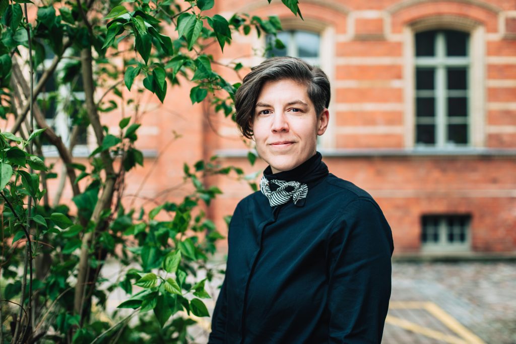 Das Bild zeigt Katrin Horn. Eine Frau mit kurzen, braunen Haaren, einem dunklen Oberteil vor einem Backsteingebäude.
