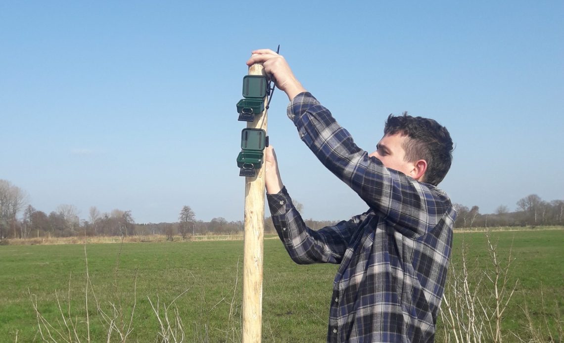 Auf dem Foto ist Alexander Drexler zu sehen. Er montiert gerade ein Gerät für die Biodiversitätserfassung per Audio an einem Holzpfahl. Im Hintergrund ist eine große Grünfläche zu sehen.