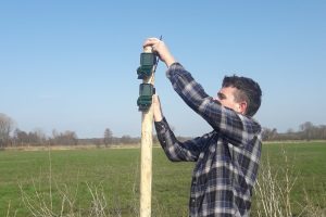 Auf dem Foto ist Alexander Drexler zu sehen. Er montiert gerade ein Gerät für die Biodiversitätserfassung per Audio an einem Holzpfahl. Im Hintergrund ist eine große Grünfläche zu sehen.