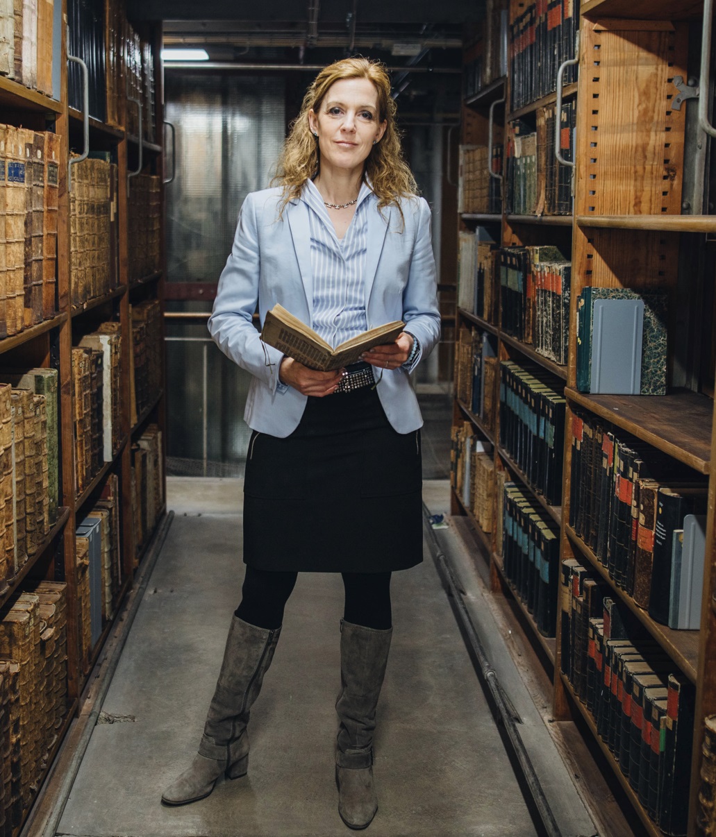 Auf dem Foto ist eine blonde Frau zu sehen, die zwischen zwei Regalen in einem Archiv steht und ein historisches Buch in den Händen hält. Sie lächelt freundlich in die Kamera.