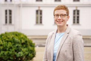 Das Foto zeigt die Junior-Professorin Dr. Marisa Holzapfel. Sie hat kurze, rotblonde Haare, trägt eine Brille und lächelt sehr freundlich in die Kamera. Im Hintergrund ist das Gebäude der Hochschulkommunikation der Uni Greifswald zu sehen.