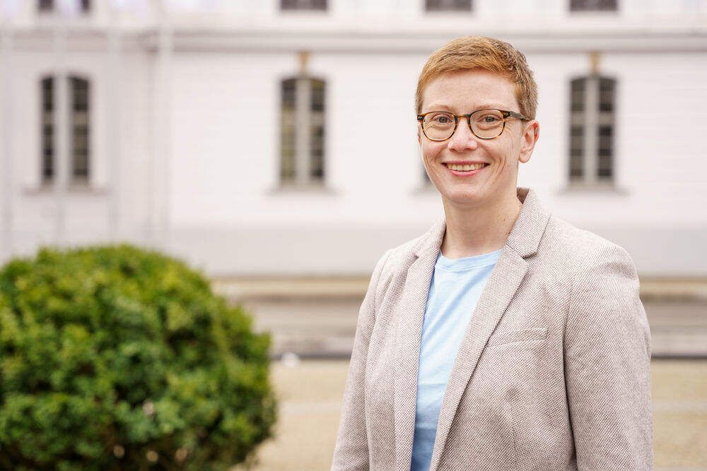 Das Foto zeigt die Junior-Professorin Dr. Marisa Holzapfel. Sie hat kurze, rotblonde Haare, trägt eine Brille und lächelt sehr freundlich in die Kamera. Im Hintergrund ist das Gebäude der Hochschulkommunikation der Uni Greifswald zu sehen.