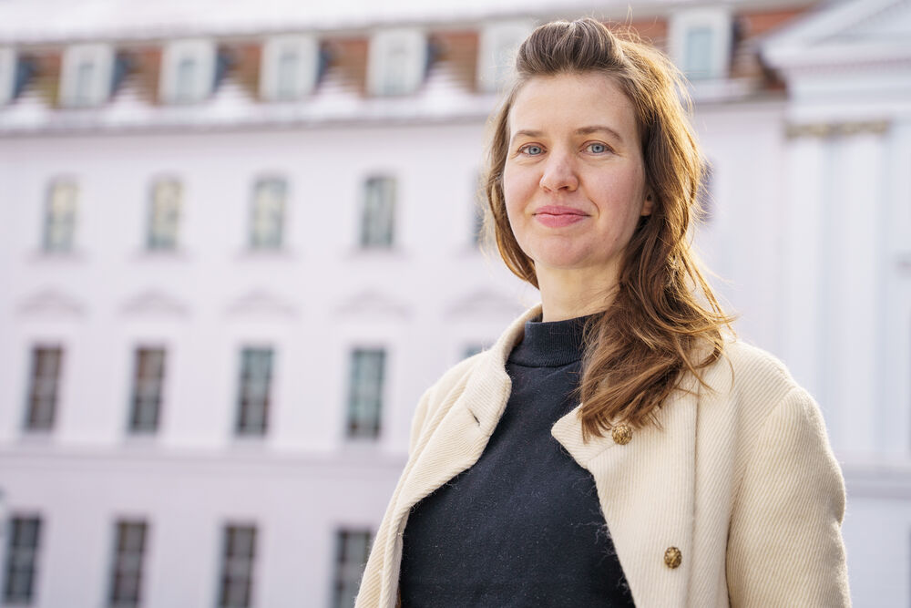 Das Bild zeigt die Junior-Professorin Dr. Elisabeth Flucher. Sie hat lange braune Haare und lächelt freundlich in die Kamera. Im Hintergrund ist das Gebäude der Hochschulkommunikation zu sehen.