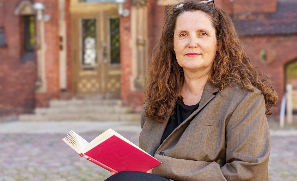 Das Foto zeigt Prof. Dr. Heide Volkening. Sie hat lange, gelockte braune Haare, die von einer Brille auf ihrem Kopf zurückgehalten werden. In ihrem Schoß hat sie ein aufgeschlagenes, rotes Buch und sie lächelt freundlich in die Kamera.