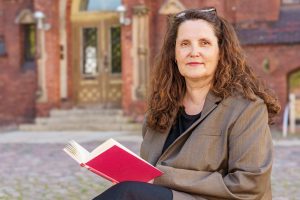 Das Foto zeigt Prof. Dr. Heide Volkening. Sie hat lange, gelockte braune Haare, die von einer Brille auf ihrem Kopf zurückgehalten werden. In ihrem Schoß hat sie ein aufgeschlagenes, rotes Buch und sie lächelt freundlich in die Kamera.