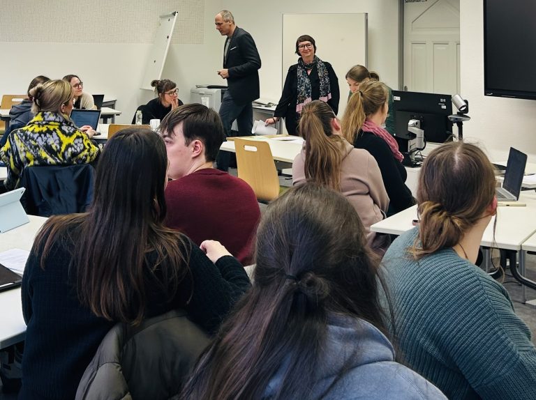 Auf dem Foto ist ein gut gefüllter Seminarraum zu sehen. Die Studierenden sitzen an Gruppentischen und hören gerade der Dozentin zu, die vorne im Raum etwas erklärt.