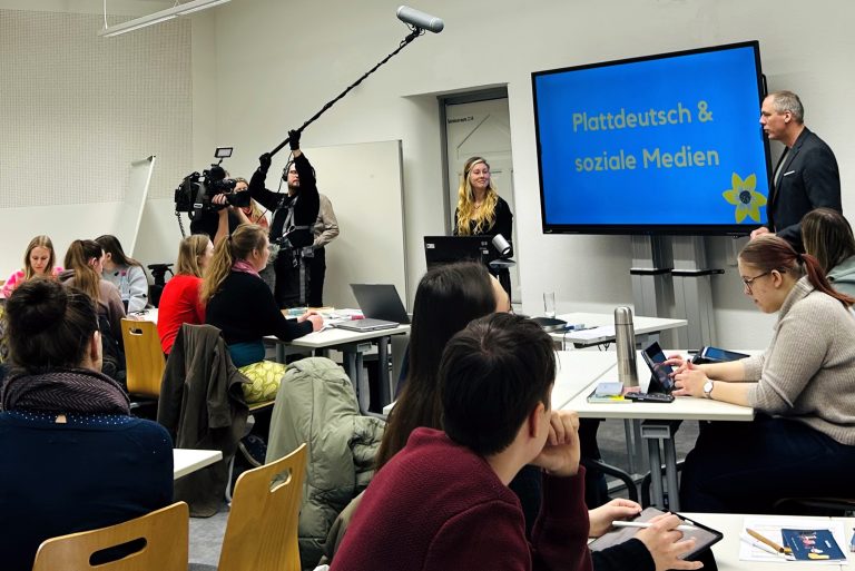 Das Foto zeigt einen Workshop zum Thema „Plattdeutsch & Social Media“. Die Studierenden sitzen an Gruppentischen und schauen nach Vorne in Richtung Bildschirm an der Tafel. Auf der linken Seite des Raums zeichnet ein Kamera-Team mit Mikrofon den Workshop auf.