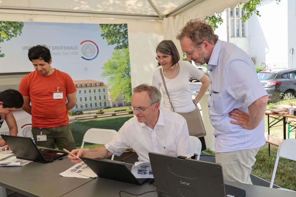 Ein Mann sitzt an einem Computer, zwei Personen schauen ihm dabei zu.