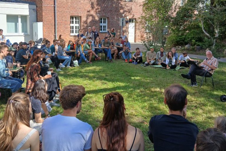 Das Bild zeigt den Garten eines Hauses, in dem ein großer Sitzkreis von Studierenden gebildet wurde. Sie hören Hans Joosten zu, der auf einem Stuhl in dem Sitzkreis sitzt.