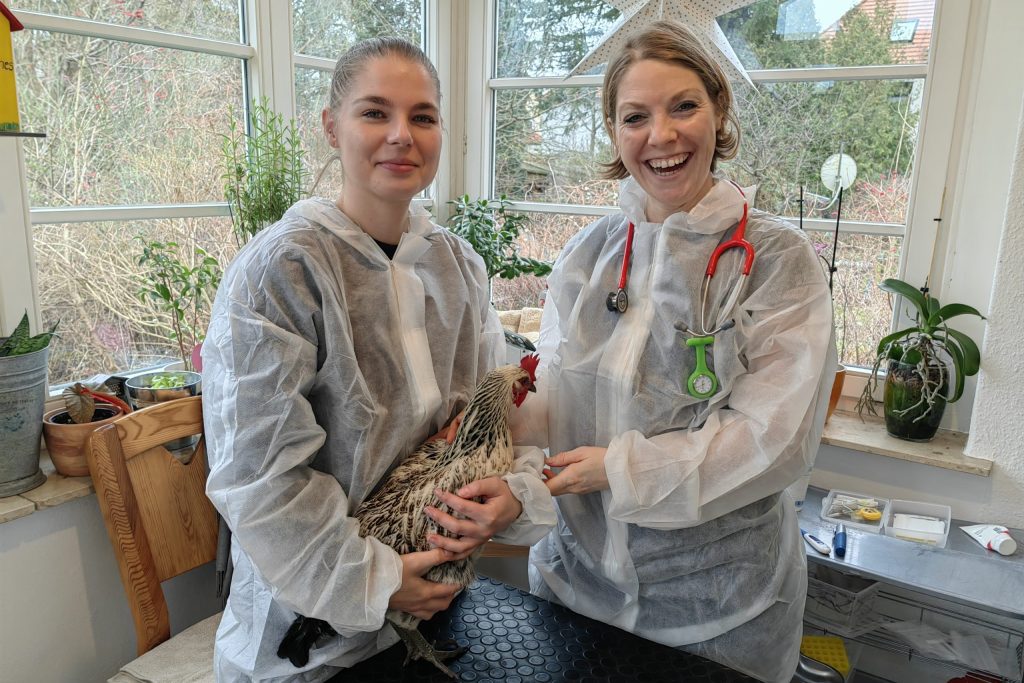 Das Foto zeigt zwei Frauen in durchsichtigen Schutzkitteln. Die Frau links im Bild hält ein Huhn im Arm, die rechte Frau, die ein Stethoskop um den Hals hängen hat, hat ihren Arm freundlich um die jüngere Frau links im Bild. Beide Frauen lächeln freundlich in die Kamera.
