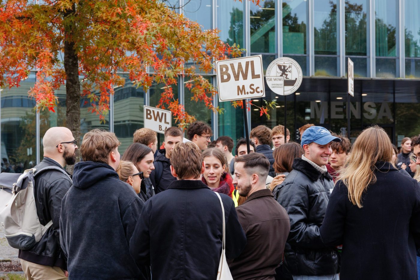Mehrere Menschen reden miteinander bei der Begrüßung in der Ersti-Woche, während die Tutor*innen Schilder mit den jeweiligen Fächern hochhalten.