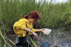 The photo shows Mariia Leontieva on an excursion in Poland. She stands dressed in a waterproof trouser suit in a river and holds a landing net in her hand.