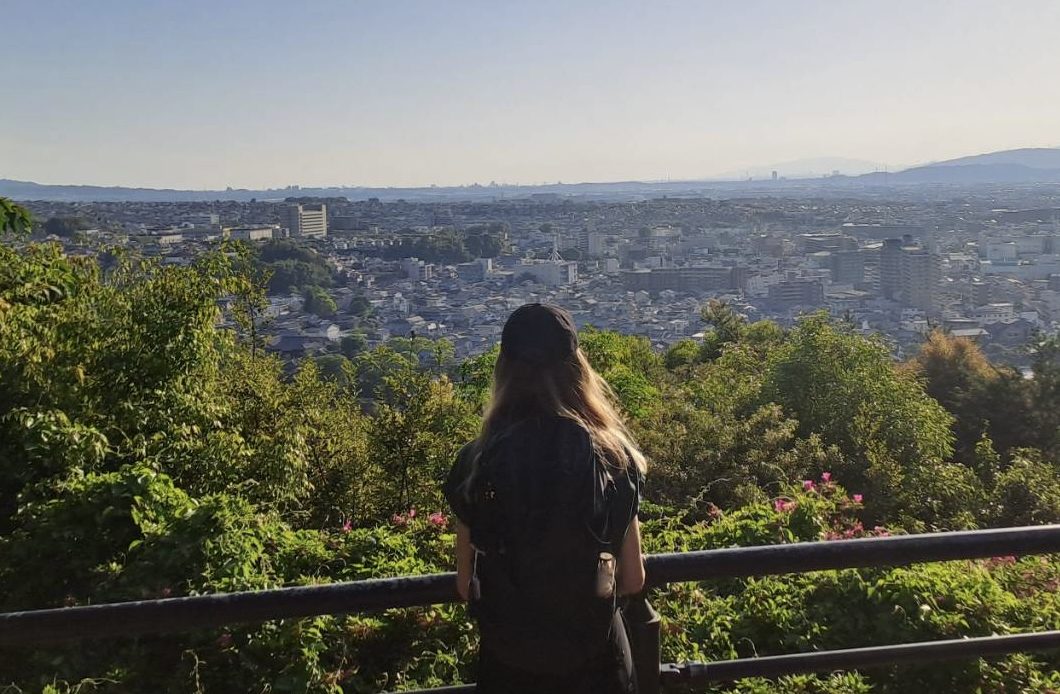 Das Foto zeigt Lisa Bähr, wie sie von einem Aussichtspunkt über die Stadt Uji (Japan) schaut. Sie wird von hinten fotografiert, weshalb ihr Gesicht nicht zu sehen ist. Lisa trägt ein dunkles T-Shirt, einen Rucksack und Basecap auf ihren langen Haaren.