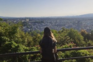 Das Foto zeigt Lisa Bähr, wie sie von einem Aussichtspunkt über die Stadt Uji (Japan) schaut. Sie wird von hinten fotografiert, weshalb ihr Gesicht nicht zu sehen ist. Lisa trägt ein dunkles T-Shirt, einen Rucksack und Basecap auf ihren langen Haaren.
