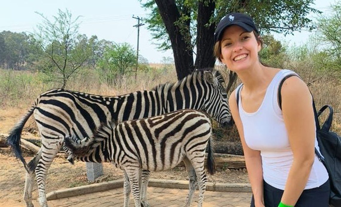 Das Foto zeigt Leona, die breit in die Kamera lächelt. Sie hat ihre Haare zu einem Zopf gebunden und trägt ein Basecap. Im Hintergrund ist ein Zebra mit seinem Kalb zu sehen, das gerade bei der Mutter trinkt.