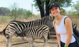Das Foto zeigt Leona, die breit in die Kamera lächelt. Sie hat ihre Haare zu einem Zopf gebunden und trägt ein Basecap. Im Hintergrund ist ein Zebra mit seinem Kalb zu sehen, das gerade bei der Mutter trinkt.