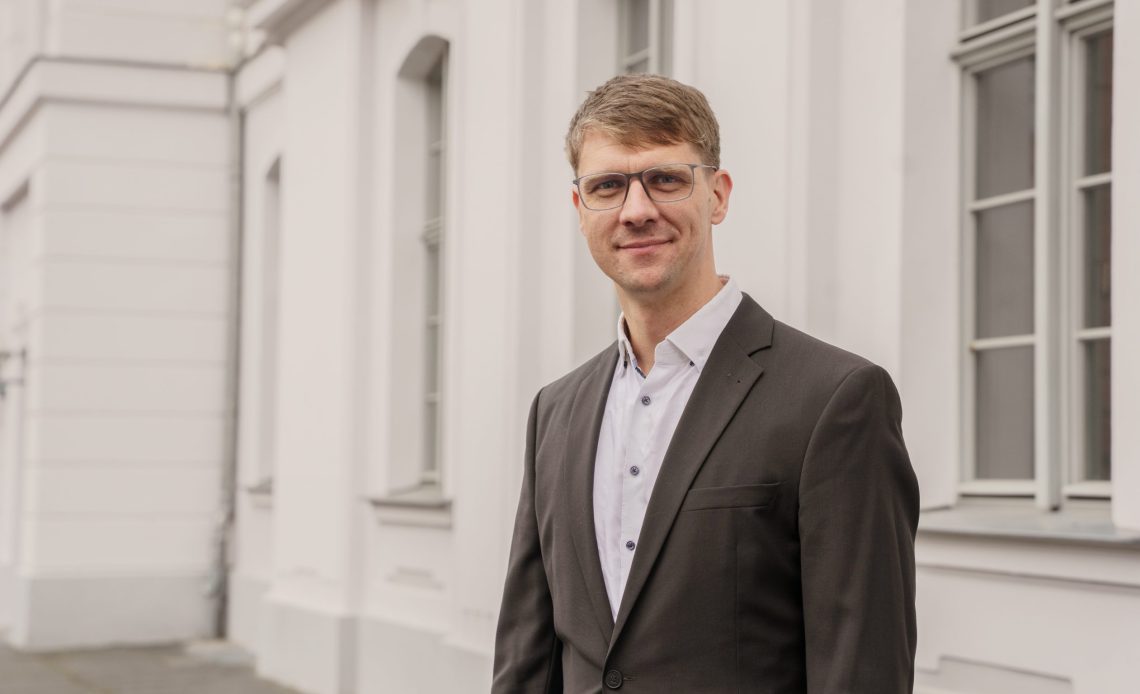 Das Foto zeigt Prof. Dr. Hendrik Lohse-Bossenz. Er trägt einen dunkelgrauen Anzug, hat kurze braune Haare und trägt eine Brille. Er lächelt freundlich in die Kamera.