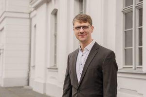 Das Foto zeigt Prof. Dr. Hendrik Lohse-Bossenz. Er trägt einen dunkelgrauen Anzug, hat kurze braune Haare und trägt eine Brille. Er lächelt freundlich in die Kamera.