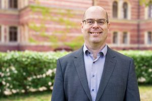 Das Bild zeigt Martin Siegel vor einem historischen Gebäude. Er trägt einen Anzug, hat eine rechteckige Brille und lächelt freundlich in die Kamera.