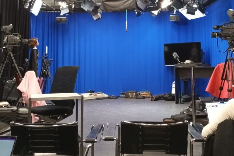Das Foto zeigt ein Studio mit verschiedenen Beleuchtungen an der Decke, einem blauen Vorhang an der Wand und Stühlen im Vordergrund des Bildes.