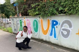 Das Fotot zeigt Jeanne D’Arc neben einer Mauer auf der in bunten Buchtaben „Fribourg“ geschrieben steht. Jeanne D’Arc Kniet neben den Buchstaben und und lächelt breit in die Kamera.