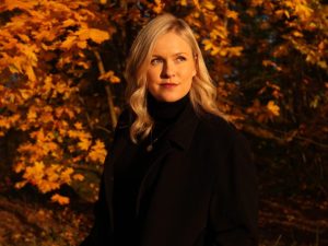 Eine Frau mit blonden Haaren steht im Herbst vor Bäumen, welche bräunliche Blätter haben