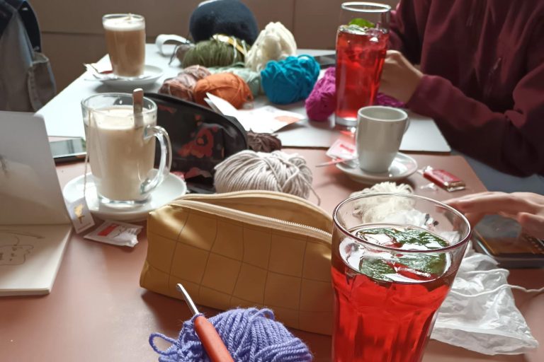 Das Bild zeigt einen Tisch in einem Café, auf dem verschiedene Wollknäuel liegen, sowie Häkelnadeln und Getränke.