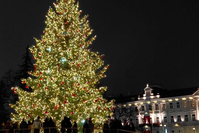 Es ist ein riesiger Weihnachtsbaum auf einem öffentlichen Platz zu sehen, der bunt geschmückt und hell beleuchtet ist. Um den Baum stehen Menschen, die Fotos machen und im Hintergrund ist ein beleuchtetes historisches Gebäude zu sehen.
