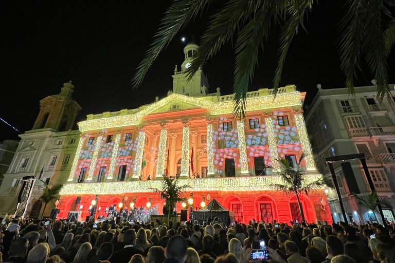 Das Bild zeigt ein bunt beleuchtetes historisches Gebäude, das in vielen hellen Farben wie geld, rosa und orange angestrahlt ist. Vor dem Gebäude ist eine große Menschenansammlung zu sehen.