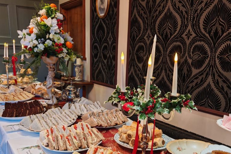 Es ist ein festlich gedeckter Tisch zu sehen, mit Blumensträußen und Kerzenhaltern. Auf dem Tisch stehen Sandwiches und verschiedene andere traditionelle britische Häppchen.