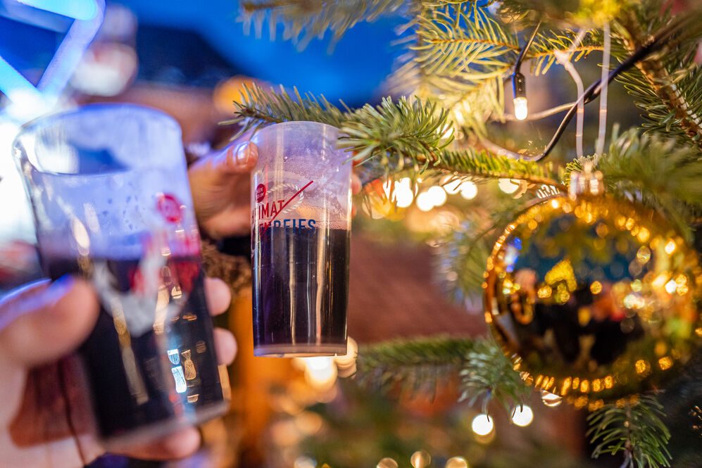 Auf dem Foto sind zwei Glühweinbecher zu sehen, die neben einen geschmückten Tannenbaum gehalten werden. Im Hintergrund ist bereits dunkel, wodurch die goldene Kugel des Tannenbaums und die leuchtenden Lichter besonders hervorstechen.