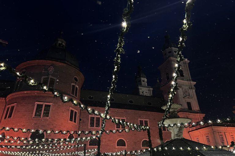 Das Foto zeigt Lichterketten, die den gesamten Weihnachtsmarkt in Salzburg beleuchten. Im Hintergrund ist ein Teil des Doms zu sehen, der rosa angestrahlt ist
