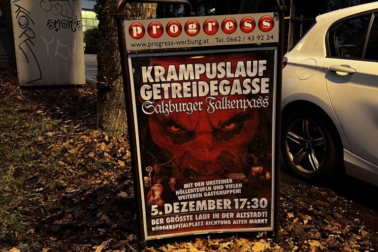 Es ist ein Schild mit einem großen Werbeplakat zu sehen, auf dem zum Krampuslauf in der Salzburger Altstadt informiert wird. Auf dem Plakat ist außerdem ein rotes, teufelsähnliches Gesicht zu sehen, was einen Krampus darstellen soll.