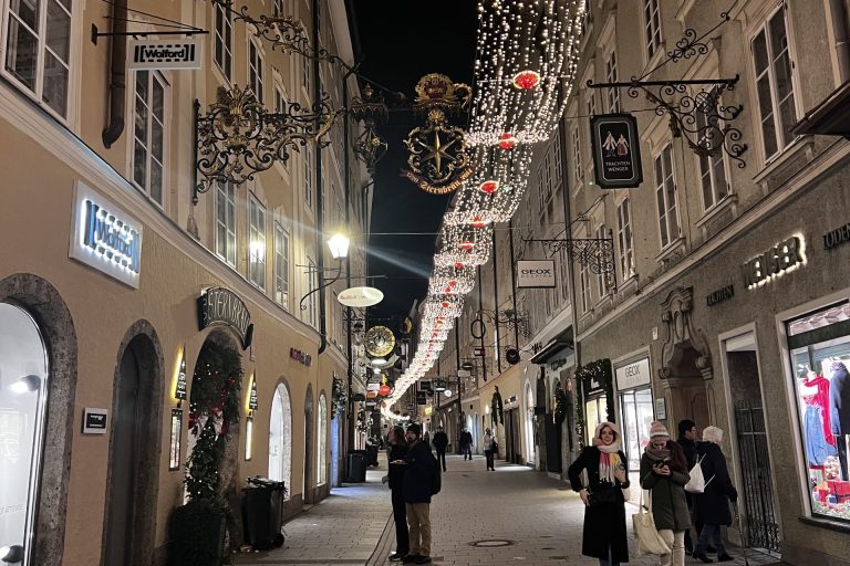 Das Foto zeigt die Getreidegasse, die wohl bekannteste Einkaufsstraße in Salzburg. Es ist dunkel, wodurch der weihnachtliche Schmuck, der in der Gasse angebracht wurde, in hell-weißem und roten Licht leuchtet.