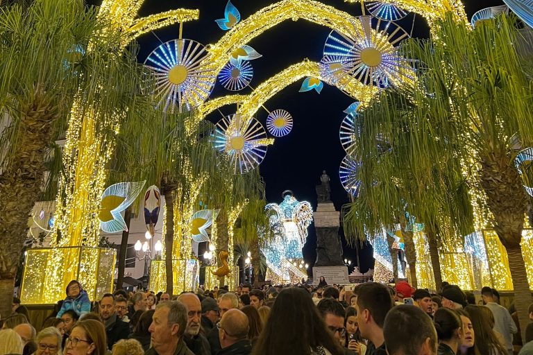 Es ist eine Art Allee zu sehen, über die sich bunte Lichtbögen und weiterer bunt leuchtender Schmuck biegen. Im Vordergrund sind viele Menschen zu sehen, die durch die Straße laufen.