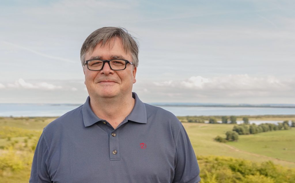 Prof. Dr. Marko Pantermöller steht auf einem kleinen Hügel, im Hintergrund sind Wiese, Bäume und Wasser zu sehen