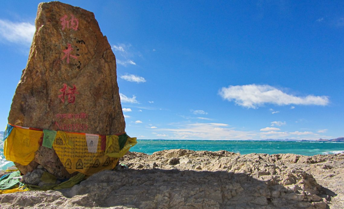 Blick auf den blauen Nam Co-Salzsee und einen Gebetsstein, der mit Inschriften versehen ist.