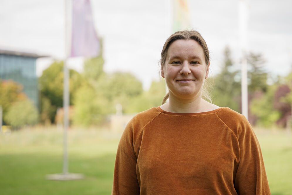 Das Foto zeigt Prof. Dr. Josephine Thomas. Sie hat braune Haare, die sie im Nacken zu einem Zopf zusammengebunden hat, trägt ein orangenes langes Oberteil und lächelt freundlich in die Kamera. Im Hintergrund sind wehende Fahnen auf einer grünen Wiese zu sehen.