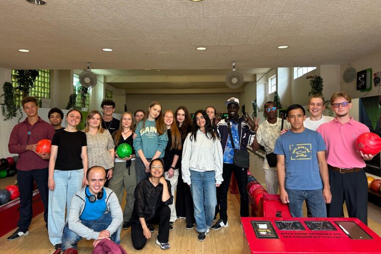 Auf dem Foto sieht man die Gruppe in einer Bowlinghalle mit Bowlingkugeln in der Hand. Sie lächeln in die Kamera.