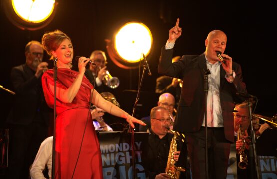 Eine Frau und ein Mann, die schick gekleidet sind, stehen auf einer Bühne mit einer Big Band und halten Mirkos in die sie reinsingen.