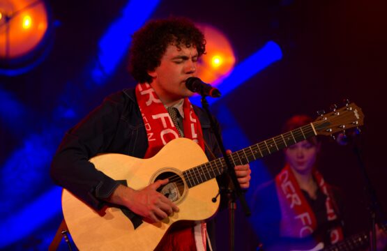 Ein junger Mann steht mit Gitarre und singt in ein Mikrofon.