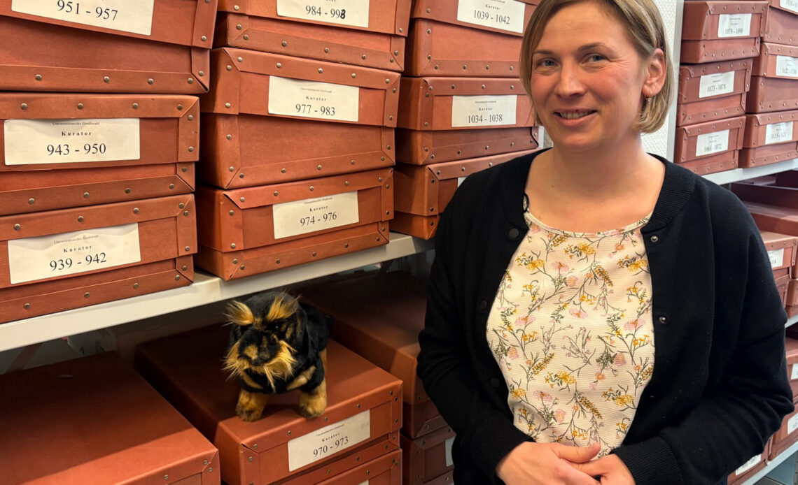 Eine Frau mit Bluse mit Blumenmuster steht vor vielen orangefarbenen beschrifteten Regalkästen.