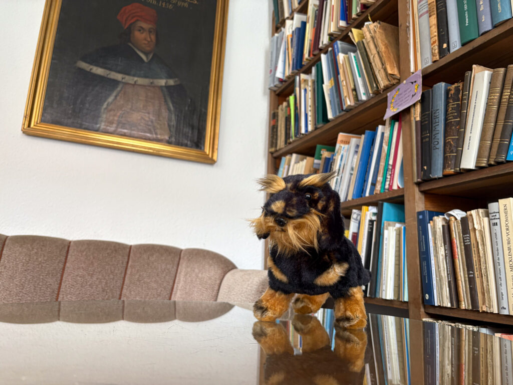 Ein Plüsch-Hund sitzt auf einem Sofa in einem Raum mit Büchern und einem Gemälde an der Wand.