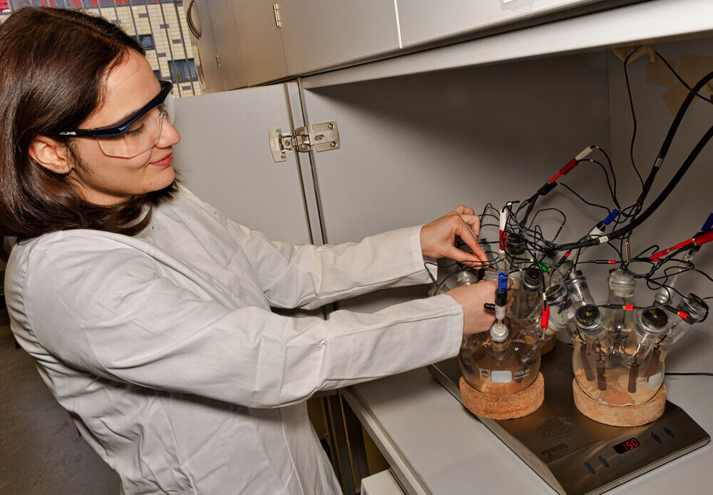 Auf dem Foto ist eine braunhaarige Frau zu sehen, die eine Schutzbrille und einen Laborkittel trägt. Sie arbeitet an kompliziert aussehenden Konstrukten aus Glas und Kabeln.
