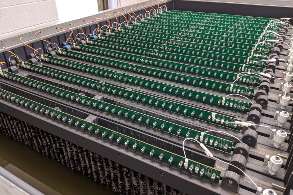 Das Foto zeigt ein sehr großes Konstrukt aus zahlreichen grauen Reihen, an denen unten längliche Bürstchen angebracht sind. Auf diesen Reihen sind grüne lange Platten angebracht, an denen Technik installiert ist. An den Seiten sind weitere Kabel angebracht.