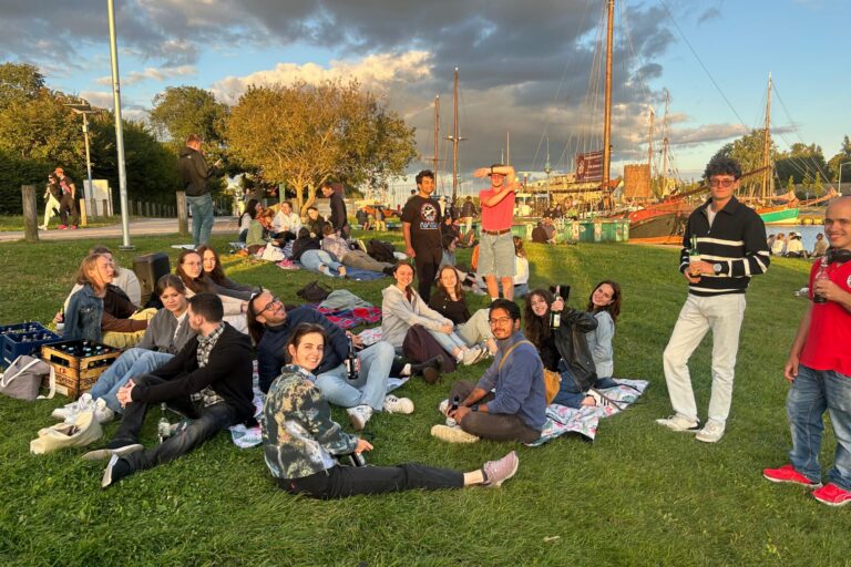 Auf dem Foto sitzen die Teilnehmenden auf der Wiese am Greifswalder Hafen. Es ist dämmriges Licht, die Sonne geht gerade unter und im Hintergrund sind einige im Hafen liegende Segelboote zu sehen.