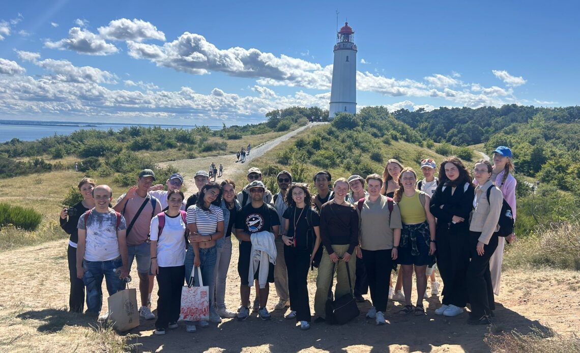 Das Foto zeigt die Gruppe der Teilnehmer*innen des Greifswald Summer. Sie stehen vor einem Leuchtturm auf Hiddensee und lächeln freundlich in die Kamera.